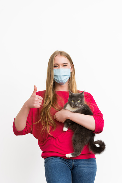 joven mujer en médico máscara celebración en brazos lindo gato y mostrando pulgar hacia arriba aislado en blanco
  - Foto, imagen