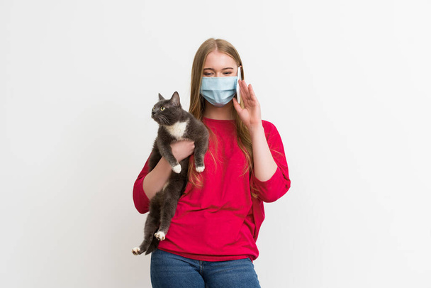 jovem mulher em máscara médica segurando nos braços bonito gato e falando no smartphone isolado no branco
  - Foto, Imagem
