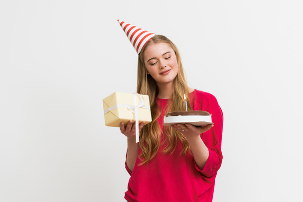 glückliches Mädchen in Partymütze schaut auf Geburtstagstorte und hält Geschenk isoliert auf weiß  - Foto, Bild