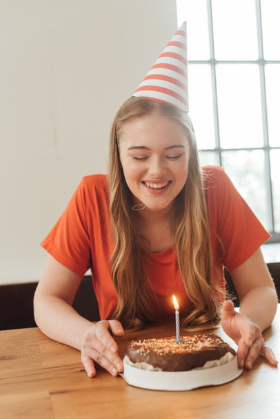cheerful woman in party cap looking at delicious birthday cake  - Фото, изображение