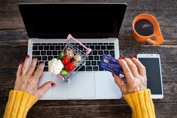 Oudere vrouw maakt online winkelen met creditcard op tafel. E commerce concept met winkelwagen vooral na coronavirus. - Foto, afbeelding