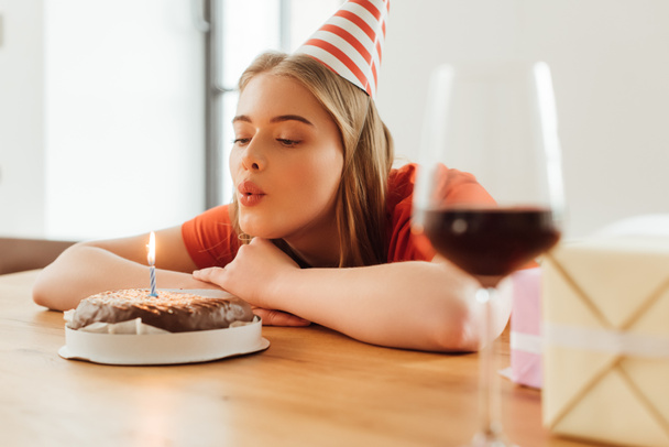 selective focus of girl in party cap blowing out candle on birthday cake near presents and wine glass on table - Photo, Image