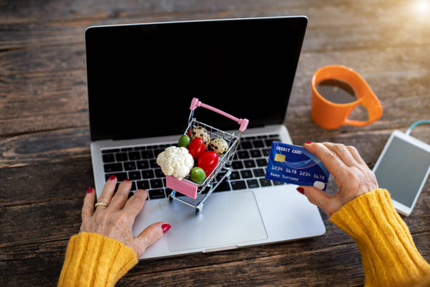 Mujer mayor hace compras en línea con tarjeta de crédito sobre la mesa. Concepto de comercio electrónico con carrito de compras, especialmente después de coronavirus
. - Foto, Imagen