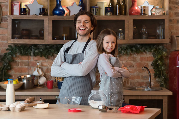 Valmiina leivontaan. Isä ja pieni tytär poseeraa keittiössä yllään esiliinat
 - Valokuva, kuva