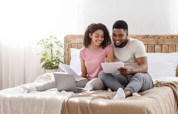 Joven pareja negra sentada en la cama en casa, comprobando el presupuesto
 - Foto, imagen