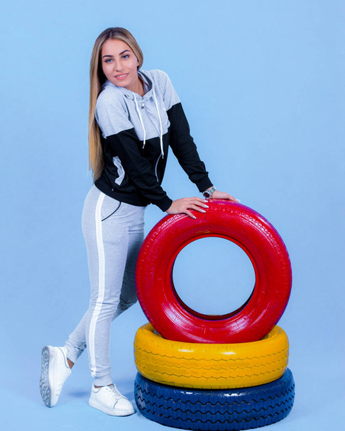 pretty young blonde model girl in sportswear posing next to car multi-colored tires in blue backround, - Photo, Image