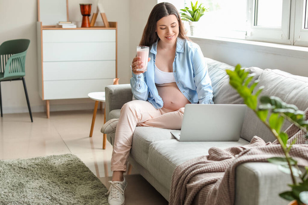 Mulher grávida bonita com smoothie e laptop em casa
 - Foto, Imagem