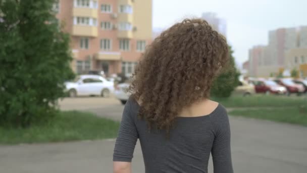 back view. woman with curly hair walks along a city street, the wind in her hair - Materiał filmowy, wideo
