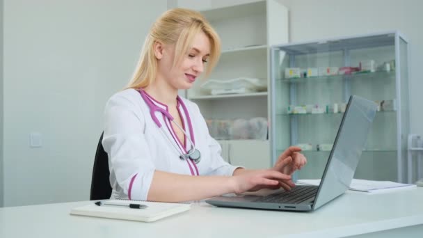 Gelukkige jonge vrouw arts in het ziekenhuis in witte jas met behulp van laptop computer schrijven notities op de werkplek. Medic doet online onderzoek, denkt na over uitdaging of probleemoplossing concept - Video