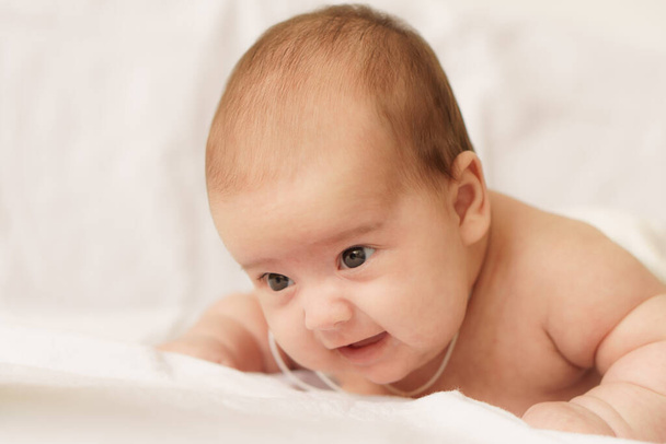 Little newborn baby in a white bodysuit lays on stomach on the bed. Top view of a newborn baby on a white warm blanket. The newborn is awake looking around indoors - Photo, Image