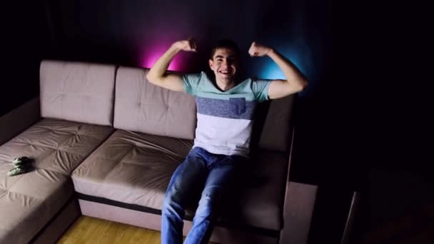 Excited young man having fun enjoy victory. Emotional and smiling guy is sitting on the couch in front of the TV. Showing biceps muscles hands. People sincere emotions, lifestyle concept. Cozy room. - Filmagem, Vídeo