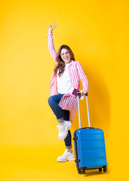 Pełne ciało zdjęcie emocjonalnie szczęśliwa dziewczyna w modnych stylowych ubrań, okulary przeciwsłoneczne, trzymając bilet i paszport, niebieska walizka korzystających rosnąca ręka pokazując znak pokoju z dwoma palcami odizolowane żółte tło - Zdjęcie, obraz