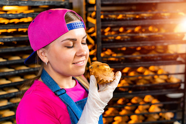 Fiatal, boldog női dolgozó steril kosztümben, frissen sült muffint tartva a bádoglemezen az élelmiszer-előállító üzemben.. - Fotó, kép