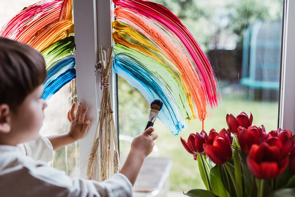 Little boy painting rainbow on a window and looking through the window while sitting home during quarantine. Let's all be well. Quarantine. Virus. Pandemic.Coronavirus. Stay home concept. Support NHS. Thank you NHS - Photo, Image