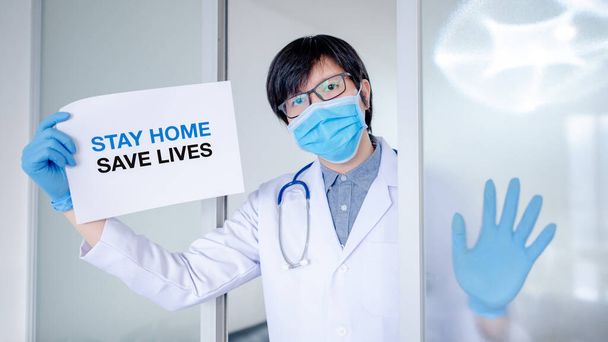 Врач или медицинский работник в защитном костюме, маске и перчатках, показывающих бумагу с сообщением Оставайтесь дома Карантинная кампания во время пандемии коронавируса (COVID-19)
. - Фото, изображение
