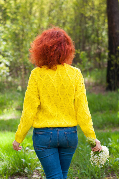 πίσω όψη κόκκινα μαλλιά κορίτσι με μπουκέτο από κρίνα της κοιλάδας σε φωτεινό κίτρινο πουλόβερ στο δάσος φόντο στην ηλιόλουστη μέρα  - Φωτογραφία, εικόνα