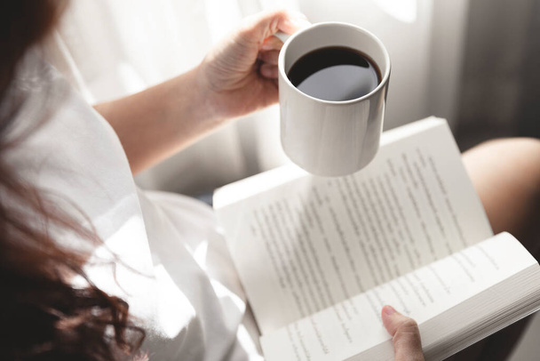 Junge Frauen, die ein Buch lesen und eine Tasse Kaffee in der Hand halten, sitzen am Fenster und ruhen sich aus, Mädchen lesen ein Buch - Foto, Bild