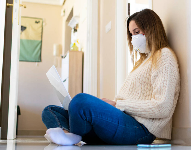 Woman sitting on the floor while teleworking from home with a mask on her face. Covid-19 quarantine. - Photo, Image
