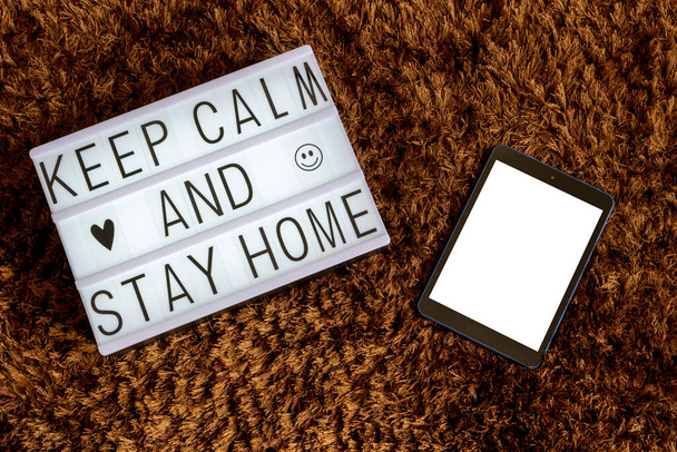 Zenithal Bild einer Leuchtbox mit einer Nachricht und einem Tablet weißen Bildschirm mit braunem Hintergrund. Coronavirus-Ratschlag: Ruhe bewahren und zu Hause bleiben. - Foto, Bild