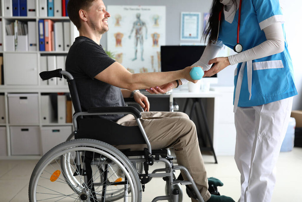 Доктор помогает человеку на инвалидной коляске держать гантель
 - Фото, изображение