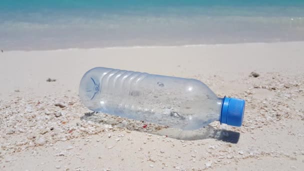 wideo z pustą plastikową butelką na białej, piaszczystej plaży i pięknymi falami w tle - Materiał filmowy, wideo