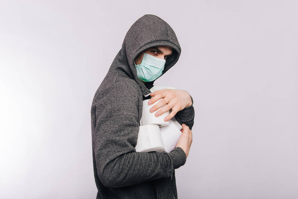 Een man in een grijze sweater en met rollen toiletpapier staat op een witte achtergrond. De opwinding tijdens quarantaine voor het coronavirus. Rotavirussen in een man met een medisch masker. - Foto, afbeelding