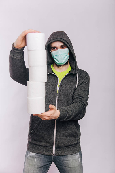 A guy in a gray sweatshirt and with rolls of toilet paper is standing on a white background. The excitement during quarantine for coronavirus. Rotaviruses in a man in a medical mask. - Photo, Image