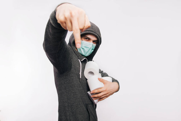 Ein Typ in grauem Sweatshirt auf weißem Hintergrund mit medizinischer Maske hält Rollen Toilettenpapier in den Händen und zeigt eine unbestimmte Geste eines Fak. Ein Mann kauft in Panik Toilettenpapier - Foto, Bild