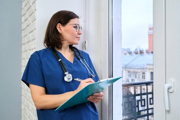 Γυναίκα νοσοκόμα με ιατρικό ιστορικό στέκεται στο γραφείο κοιτάζοντας έξω από το παράθυρο - Φωτογραφία, εικόνα