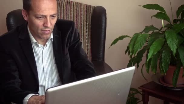 Intelligent senior looking serious working from home using his laptop with a plant on background - Кадры, видео