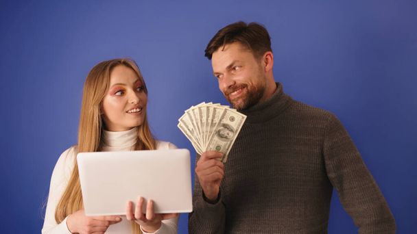Young beautiful woman showing the bill on the tablet to her rich boyfriend who holds cash in his hands. Online shopping and relationship with interest - Foto, afbeelding