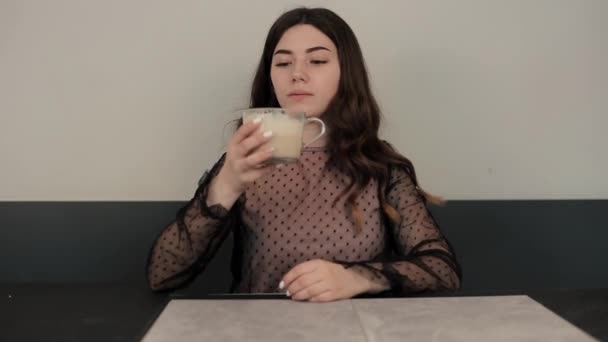 Egy vonzó fiatal lány élvezi az illatát és ízét kávé ül egy kávézóban. Egy kortyot kortyolgat, és elkezd nevetni, miközben a keze befogja a száját. Közelkép - Felvétel, videó