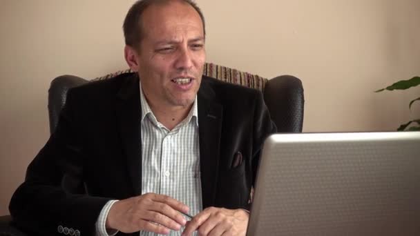 Intelligens és mosolygós idősebb üzletember távmunka otthonról kezében egy toll, amelynek videohívás kollégáival a laptopjával - Felvétel, videó