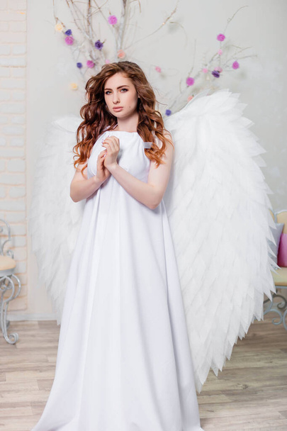 Πορτρέτο μιας γυναίκας αγγελικό κορίτσι σε ένα λευκό φόρεμα με φτερά πίσω από διπλωμένα χέρια της στην προσευχή. - Φωτογραφία, εικόνα