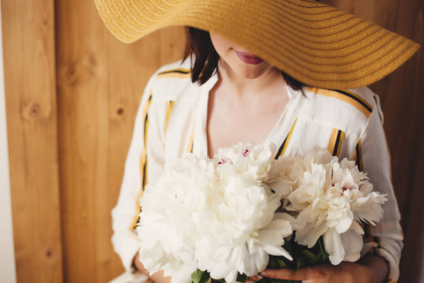 Portrait of boho woman in hat holding white peony bouquet on rustic wooden background. Stylish hipster girl in dress posing with peonies. Happy Mothers day. International Womens days - Fotoğraf, Görsel
