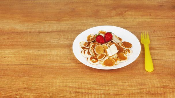 Pequeños panqueques con fresas y mantequilla están en un plato blanco sobre un fondo de madera, junto a un tenedor de plástico amarillo. Desayuno para niños, desayuno de moda, comida de moda. Espacio para texto
 - Foto, imagen