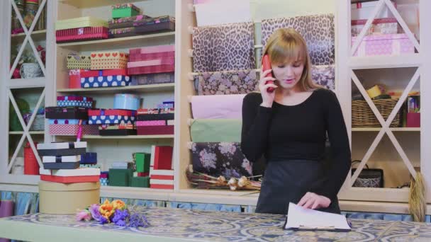 Молодая работница цветочного магазина разговаривает по телефону и проверяет в блокноте - Кадры, видео