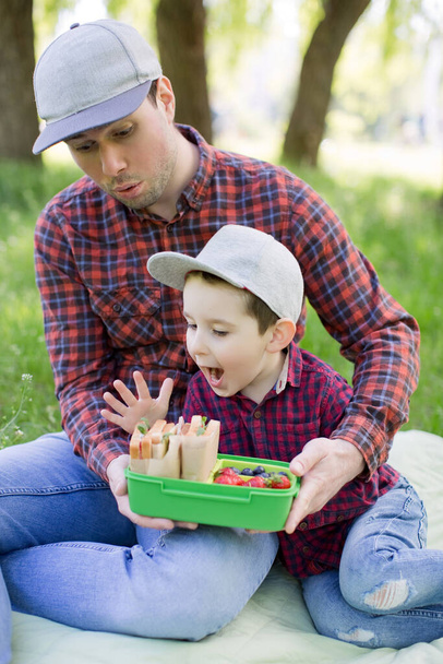 Vader en zoon eten sandwiches, aardbeien en bosbessen van groene lunchbox outdoor. Picknick in het voorjaarspark - Foto, afbeelding