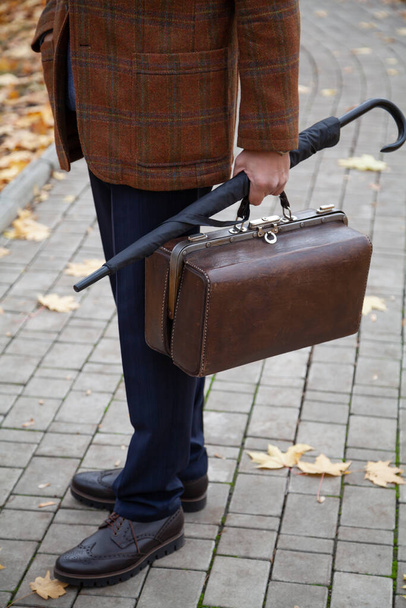 gentleman in a stylish checkered jacket stands in the alley of the square in the fall afternoon. Holding a vintage bag and umbrella cane in his han - Foto, Imagen