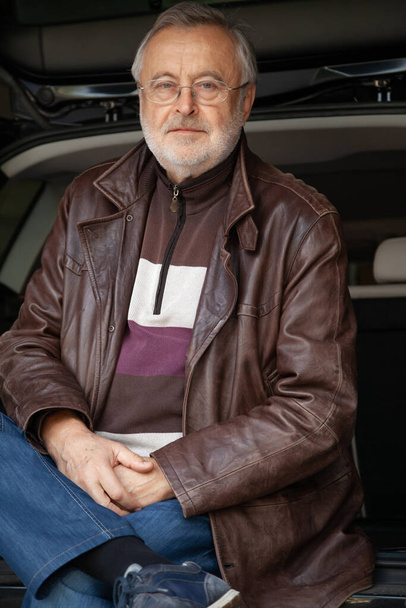 Portrait of a man with a gray beard. An elderly man in glasses and a leather jacket against the background of an open trunk of a ca - Zdjęcie, obraz