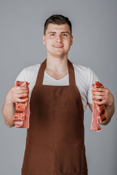 Un venditore o un cuoco maschio tiene la carne cruda tra le mani e la mostra su uno sfondo grigio. - Foto, immagini