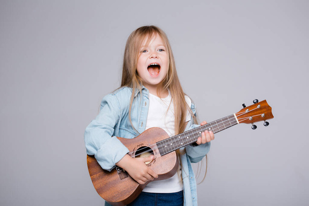 The child plays on the ukulele and sings songs. Joyful girl stands in a blue shirt on a gray background - Photo, Image