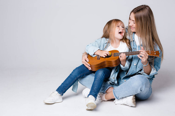 Charming Caucasian girl sings songs to her mother with a guitar. The daughter sits on the lap of the parent and enjoys the music. - Fotó, kép