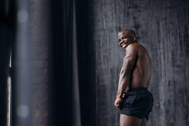 Porträt eines gut aussehenden muskulösen Afroamerikaners mit nacktem Oberkörper in schwarzen Shorts, der seinen Arm Bizeps zeigt. - Foto, Bild