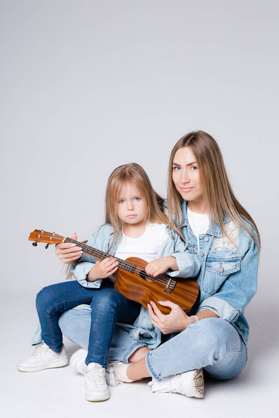A young mother and daughter of European appearance are sitting on a gray background with a guitar in their hands. Ukulele Learning - Photo, Image