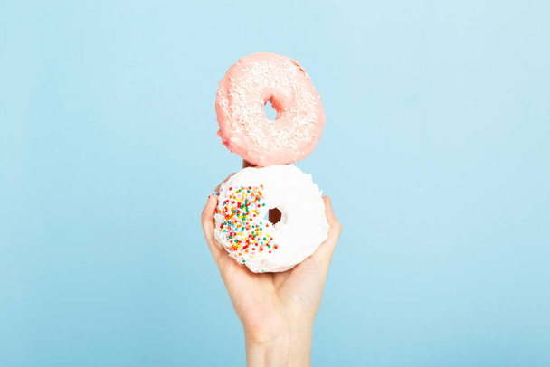 Woman hand holds two donuts with pink and white  icing and multicolored sugar pastry topping, blue background - Foto, Imagem