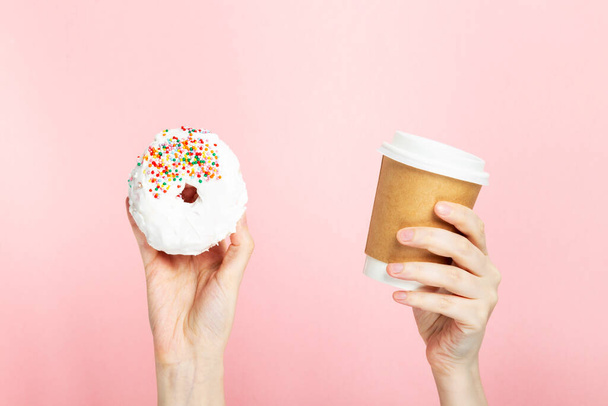Woman hand holds paper cup with coffee and donut with white icing and multicolored sugar pastry topping, pink background - 写真・画像