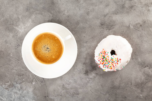 White cup of coffee and donut in white icing and multicolored sugar pastry topping on grey background. Flat lay - Foto, imagen