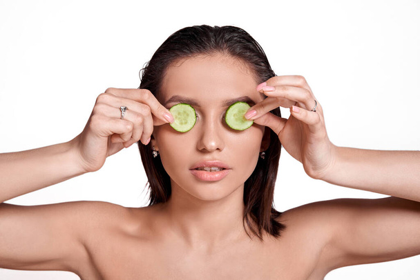 Concept Face care at home. Portrait of young beautiful woman with cucumber on her face - 写真・画像