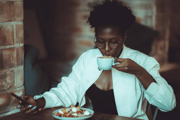 Charming elegant African businesswoman is having a brunch in a cafe with a cup of delicious cocoa and salad; young black female in eyeglasses is drinking coffee while typing a message on a cellphone - Фото, изображение